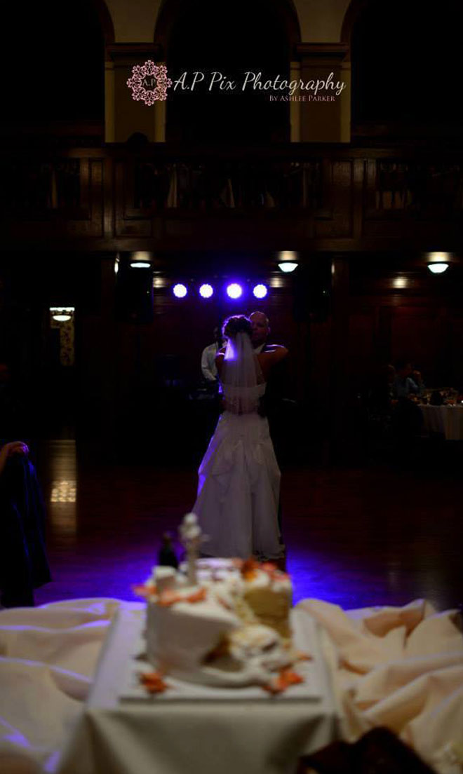Bride and Groom in first dance embrace behind wedding cake in Grand Ballroom at The Corinthian Event Center.