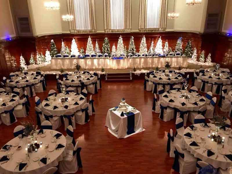 Grand Ballroom setup in white and blue theme with chair covers, flower centerpieces, and white plate place settings at The Corinthian Event Center.