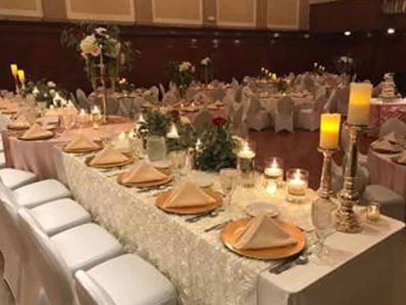 Head table with white chair covers, flower and candle centerpieces, and gold plate place settings the Grand Ballroom at The Corinthian Event Center.