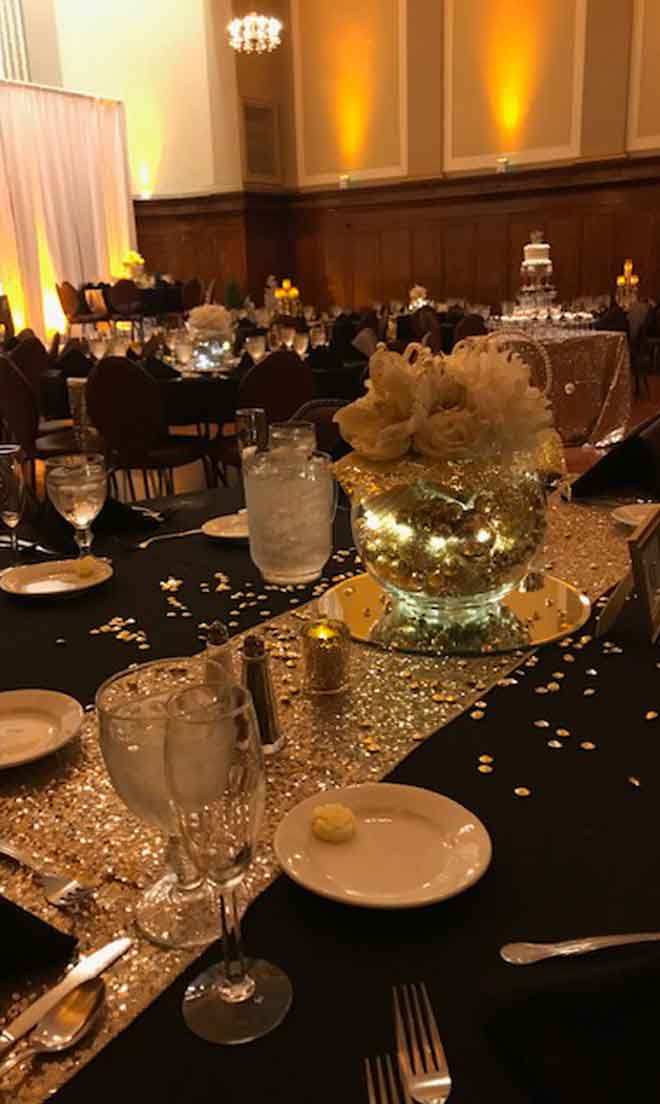 Grand Ballroom setup with stage backdrop in black theme with glittery decor centerpieces and white plate place settings at The Corinthian Event Center.