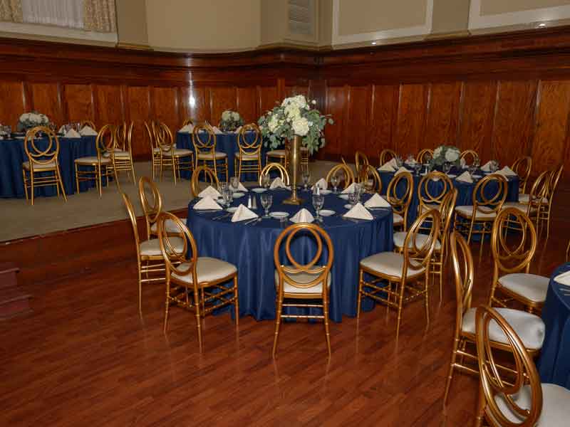 One table setup in blue and gold theme with flower centerpieces, and white place settings at The Corinthian Event Center.